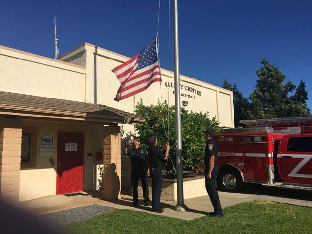 Fire station with an American flag, firefighters standing outside, and a red fire engine parked nearby.