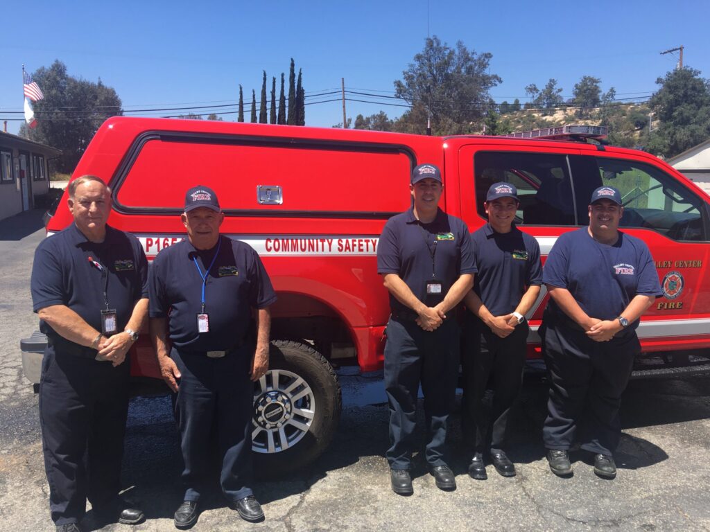 Four emergency response personnel standing in uniform beside a red fire rescue pickup truck outdoors.