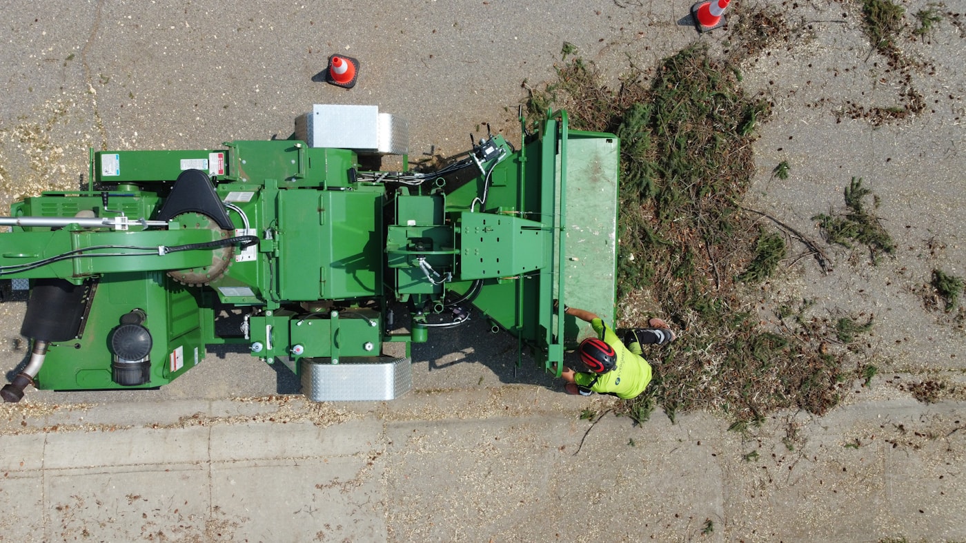 Aerial view of a green wood chipper processing a pile of brush while a worker in a yellow safety vest stands beside it.