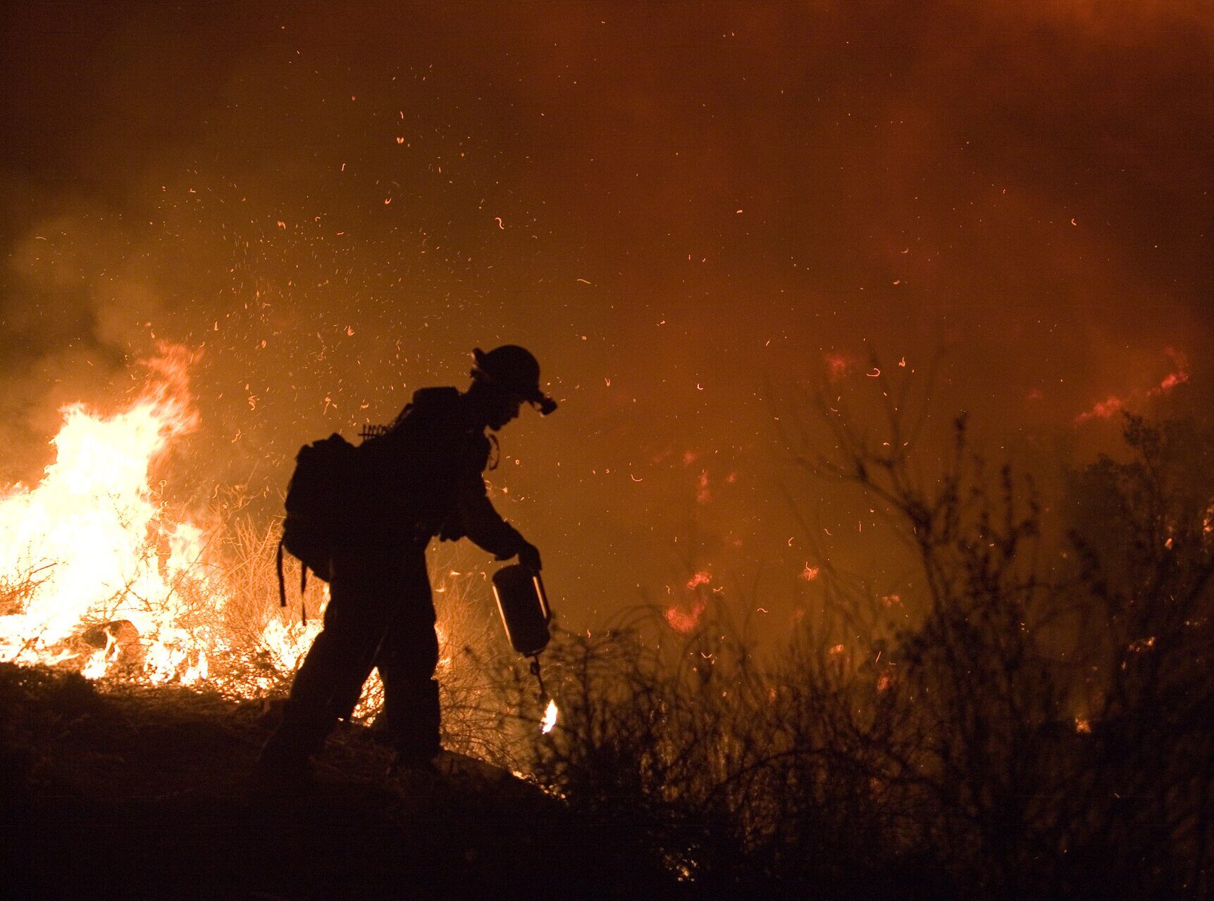 Firefighter working near flames and brush during a wildfire at night.